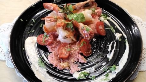 Schwarz-gelbe Garnelen liegen auf rosa Risotto, garniert mit Tomaten und Kräutern, umgeben von cremiger Soße.