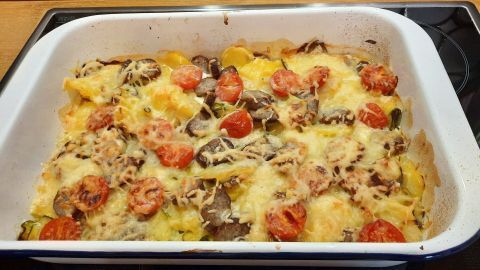 Ein Bratwurst-Gratin wird in einer weißen Auflaufform präsentiert, überbacken mit geschmolzenem Käse und Tomaten.