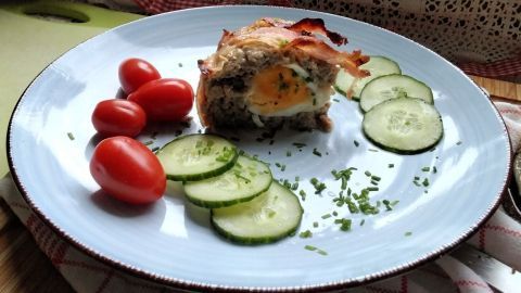 Eine saftige Hackfleischtorte mit einem offenbar weichgekochten Ei im Inneren, umgeben von Tomaten und Gurken.