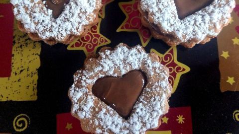 Drei Nougatplätzchen mit herzförmigen Schokoladenfüllungen und Puderzucker liegen auf einem bunten Untergrund.
