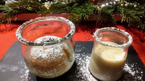 Ein Glas mit einem Bratapfel-Dessert und ein weiteres Glas mit einer cremigen Füllung stehen auf einem schwarzen Teller, umgeben von festlichem Dekor.