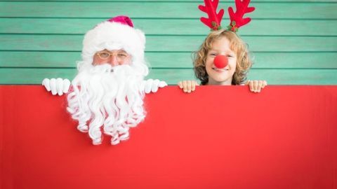 Ein Weihnachtsmann mit langer, weißer Mähne und ein Kind mit roten Rentiergeweih-Haarreif stehen hinter einem roten Schild.
