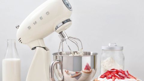 Eine stilvolle Smeg-Küchenmaschine rührt Teig, umgeben von frischen Erdbeeren, Eiern und Milch auf einem Tisch.