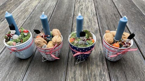 Kleine Tassen mit blauen Kerzen, gefüllt mit Nüssen und dekorativem Grün, stehen auf einem Holztisch.