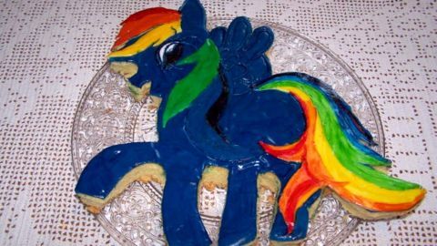 Eine farbenfrohe Motivtorte in Form von Rainbow Dash liegt auf einem gläsernen Teller und steht auf einem Tisch mit Tischdecke.