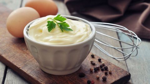 In einer Schüssel liegt selbstgemachte Mayonnaise, umgeben von Eiern und bunten Pfefferkörnern auf einem Holzbrett.