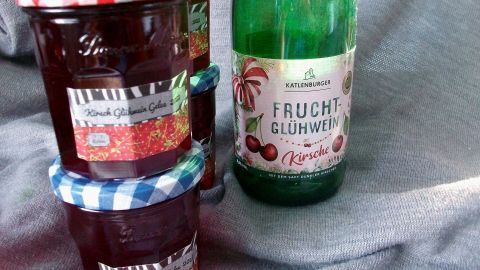 Gläser mit Kirsch-Glühwein-Gelee stehen neben einer Flasche Frucht-Glühwein, alles auf einem grauen Untergrund.