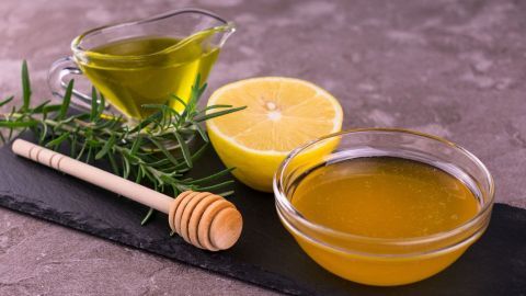 Frischer Honig und Zitronensaft liegen neben einem Olivenölkrug und Rosmarinzweigen auf einem schwarzen Steinbrett, hilfreich bei Halsschmerzen.