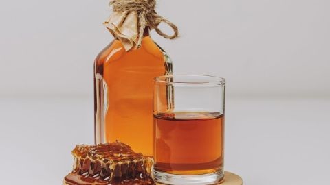 Eine Flasche mit selbstgebrautem Getränk steht neben einem Glas und einer Honigwabe auf einem Holzuntersetzer.