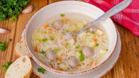 Eine Schüssel klare Suppe mit Einlagen wie Nudeln, Fleischbällchen und Gemüse steht auf einem Holztisch, umgeben von Brot und Kräutern.
