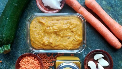 Würziger Linsenaufstrich liegt in einer Schale, umgeben von frischem Gemüse wie Karotten und Zucchini sowie Gewürzen.