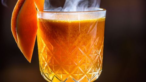 Ein dampfender Glüh-Gin in einem eleganten Glas steht auf einem Tablett mit Orangenstück und Gewürzen im Hintergrund.