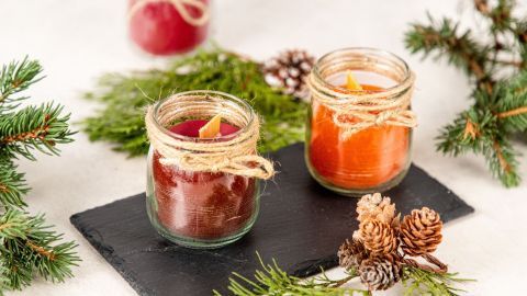 Zwei knisternde Kerzen mit Holzdochten stehen auf einer schwarzen Platte, umgeben von grünen Zweigen und Tannenzapfen.