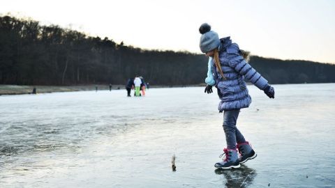 Ein Mädchen läuft vorsichtig auf dem glatten Eis eines Sees, während im Hintergrund andere Menschen das Eis genießen.