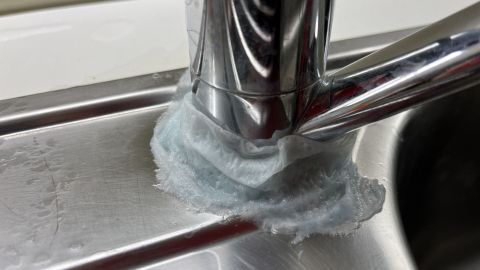 Ein glänzender Wasserhahn wird mit einer blauen Paste behandelt, während sich Wasser auf der Edelstahloberfläche sammelt.
