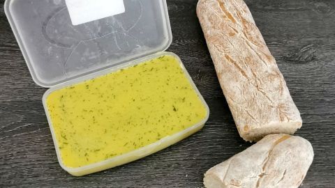 Eine Box mit Kräuterbutter liegt neben einem frisch aufgeschnittenen Brot auf einer dunklen Holzoberfläche.