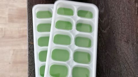 Zwei Eiswürfelformen mit grüner Flüssigkeit liegen auf einem Tisch, daneben sind bunte Silikonbecher gestapelt.