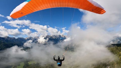 Ein Gleitschirmflieger schwebt über einer bewaldeten Berglandschaft, umgeben von Wolken und strahlend blauem Himmel.