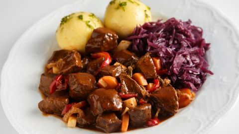 Saftiges Glühweingulasch liegt auf einem Teller, begleitet von Kartoffelknödeln und Rotkraut, in einem einladenden Ambiente.