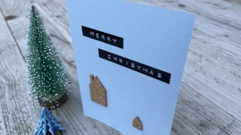 Eine Weihnachtskarte mit Korkhäuschen steht auf einem Holztisch, dekoriert mit einem kleinen, grünen Baum. "MERRY CHRISTMAS" ist auf der Karte zu lesen.