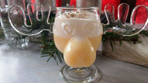Ein Glas Eierpunsch in Rentierform steht dekorativ auf einem Tisch, umgeben von Tannenzweigen und Weihnachtsdekoration.