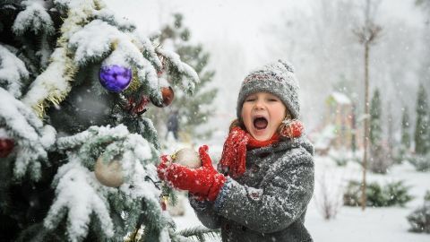 Ein Kind greift begeistert nach einer Christbaumkugel an einem geschmückten, schneebedeckten Weihnachtsbaum im Winter.