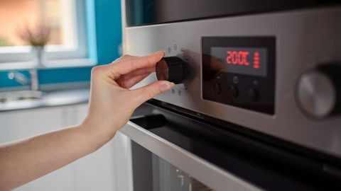 Eine Hand dreht den Regler eines modernen Backofens auf 200 Grad, um effizient zu kochen, während eine helle Küche im Hintergrund zu sehen ist.