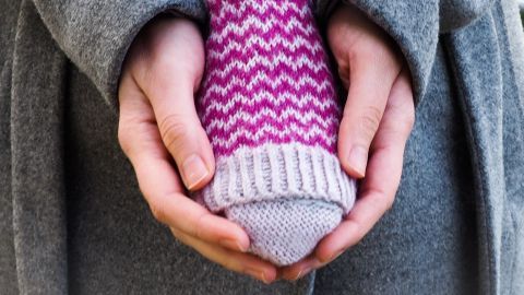 Ein gestrickter Handwärmer wird in den Händen einer Person gehalten, die einen grauen Mantel trägt, um die Kälte abzuwehren.