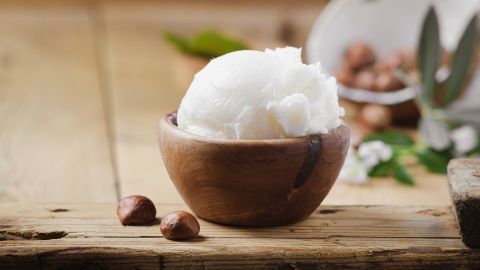 Eine Schüssel mit Shea Butter steht auf einem Holztisch, umgeben von Nüssen und grünen Blättern.
