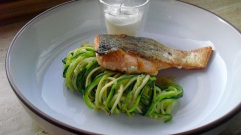 Saftiger Lachs liegt auf Zucchini-Spaghetti, neben einem kleinen Glas mit weißer Sauce, serviert auf einem weißen Teller.