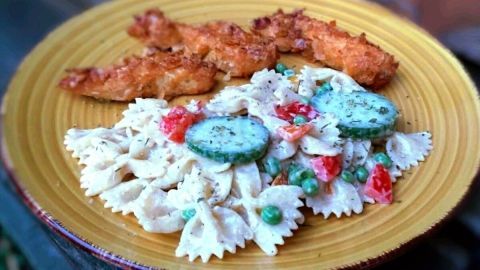 Ein bunter Nudelsalat aus Farfalle mit Erbsen, Gurken und Tomaten, serviert auf einem gelben Teller neben knusprigem Hähnchen.