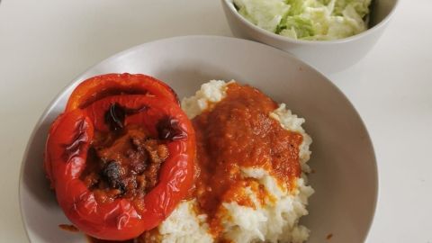 Gefüllte Paprika liegt auf einem Teller mit Reis, umgeben von einer Schüssel frischem Salat auf einem Tisch in einem hellen Raum.