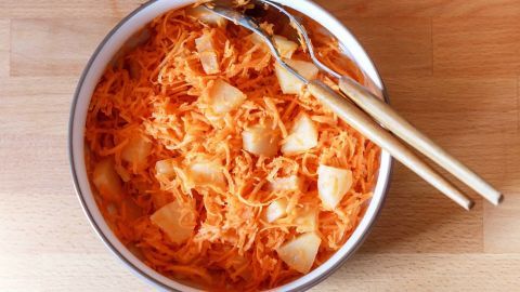 Eine Schüssel mit Karottensalat steht auf einem Holzuntergrund. Streifen aus Karotten und Äpfeln vermischen sich.