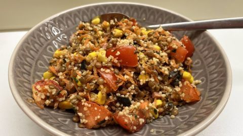 Eine Schüssel mit Couscous-Berglinsensalat enthält bunte Zutaten wie Tomaten, Mais und Linsen, angerichtet in einem dekorativen Muster.