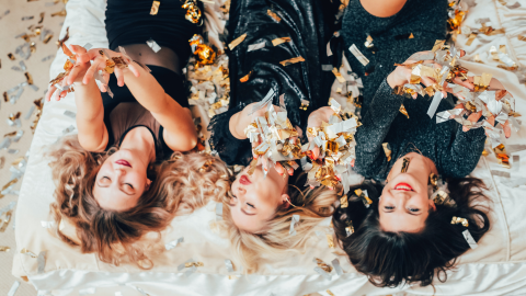 Drei Frauen in festlichen Outfits werfen Konfetti in die Luft, umgeben von einer festlichen Atmosphäre.