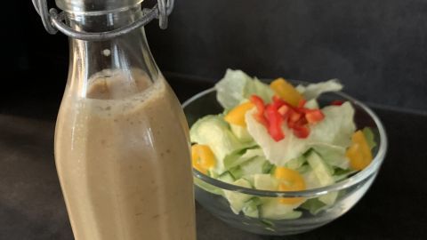 Eine Glasflasche mit leicht cremigem Dressing steht neben einer Schüssel mit frischem, buntem Salat auf einem dunklen Tisch.