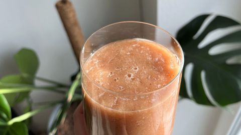Ein Glas Kombucha-Scoby-Smoothie hält jemand in der Hand, umgeben von Pflanzen in einem hellen Raum.