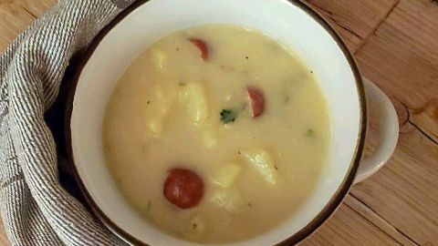 Eine cremige Kartoffelsuppe liegt in einer weißen Schüssel auf einem rustikalen Tisch, umgeben von einem grauen Geschirrtuch.