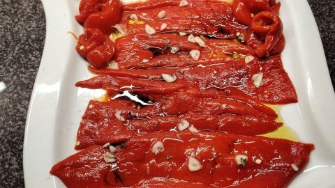 Zarte rote Paprikastreifen liegen auf einem weißen Teller, garniert mit Knoblauchstückchen und Olivenöl.