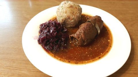 Auf einem weißen Teller liegen hausgemachte Semmelknödel, Rinderroulade und Rotkohl in würziger Soße.