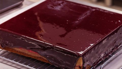 Ein quadratischer Bananen-Amaretto-Blechkuchen mit glatter Schokoladenglasur steht auf einem Gitterrost in einer Küche.