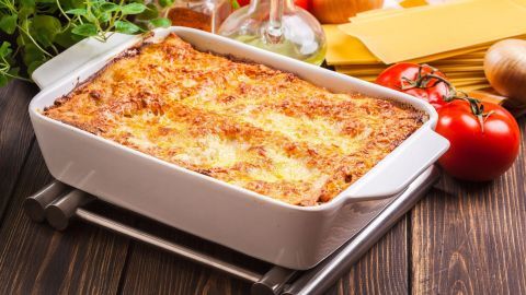 In einer weißen Auflaufform steht eine goldbraun gebackene Lasagne, umgeben von frischen Tomaten, Zwiebeln und Käse.