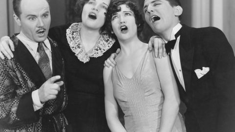 Vier Personen singen leidenschaftlich zusammen, umgeben von einem nostalgischen Inneneindruck des frühen 20. Jahrhunderts.
