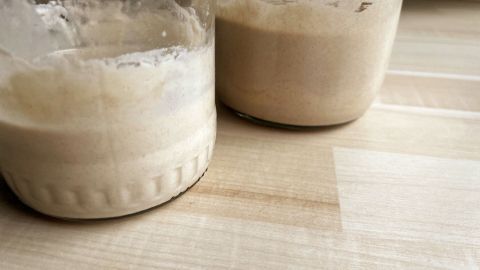 Zwei Gläser mit Roggensauerteig und Weizensauerteig stehen auf einem hellen Tisch und zeigen ihre unterschiedliche Konsistenz.