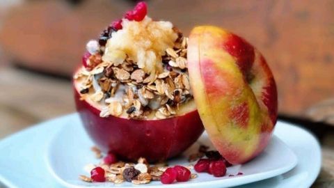 Ein gefüllter Apfel mit Früchtemüsli und Haferflocken steht auf einem weißen Teller in einer natürlichen Umgebung.