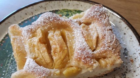 Ein Stück Apfelkuchen mit Dinkelhefeteig liegt auf einem Teller, leicht mit Puderzucker bestäubt, auf einem Tisch.