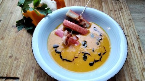 Kürbiscremesuppe mit exotischen Gewürzen liegt in einer Schüssel, dekoriert mit Speck und Kürbiskernen, auf einem Holzbrett.