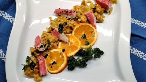 Ein weißer Teller präsentiert ein leckeres Orangen-Kürbis-Curry mit Süßkartoffel und Surimi, garniert mit frischen Kräutern.