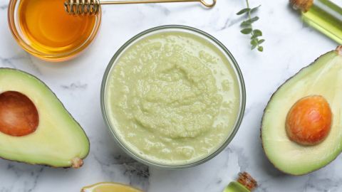 Eine grüne Gesichtsmaske aus Avocado liegt in einer Schüssel, umgeben von Honig, Zitrone und ätherischen Ölen auf Marmor.