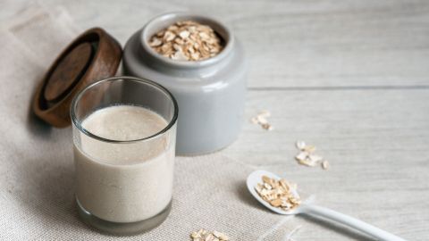 Ein Glas mit milchiger Flüssigkeit steht neben einer grauen Dose mit Haferflocken auf einem neutralen Tisch.